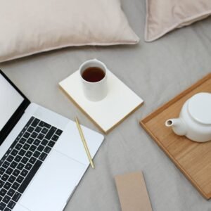 High angle of coffee cup and pot arranged on comfortable bed near opened laptop during remote work