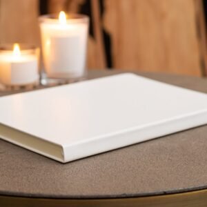 Blank stylish white notebook on round table with burning candles near wall in light room on blurred background at home